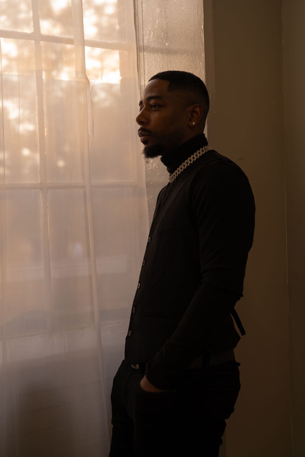 A person in a black sweater stands in profile against a window with soft sunlight filtering through sheer curtains.
