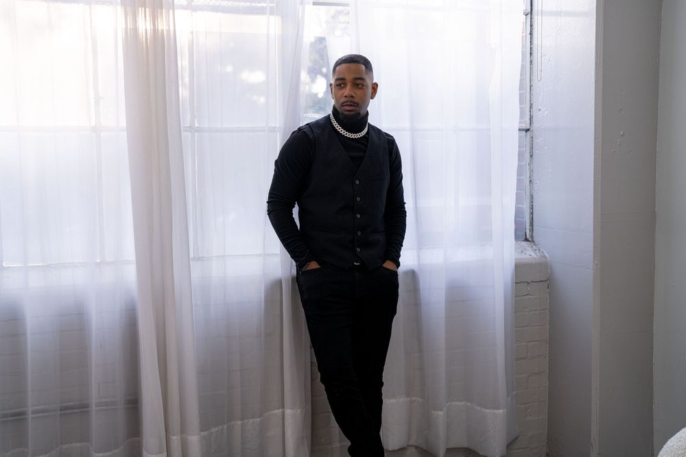 A person in black attire stands in a bright, minimalist white room with sheer curtains.