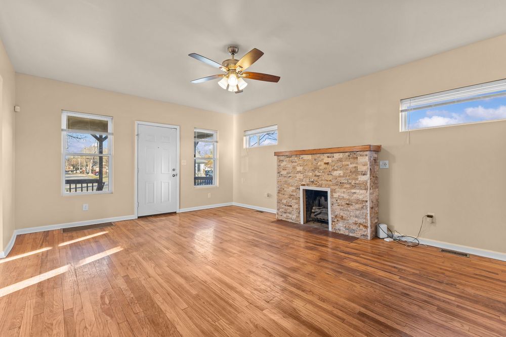 Spacious living room with hardwood floors, ceiling fan, stone fireplace, and bright windows.