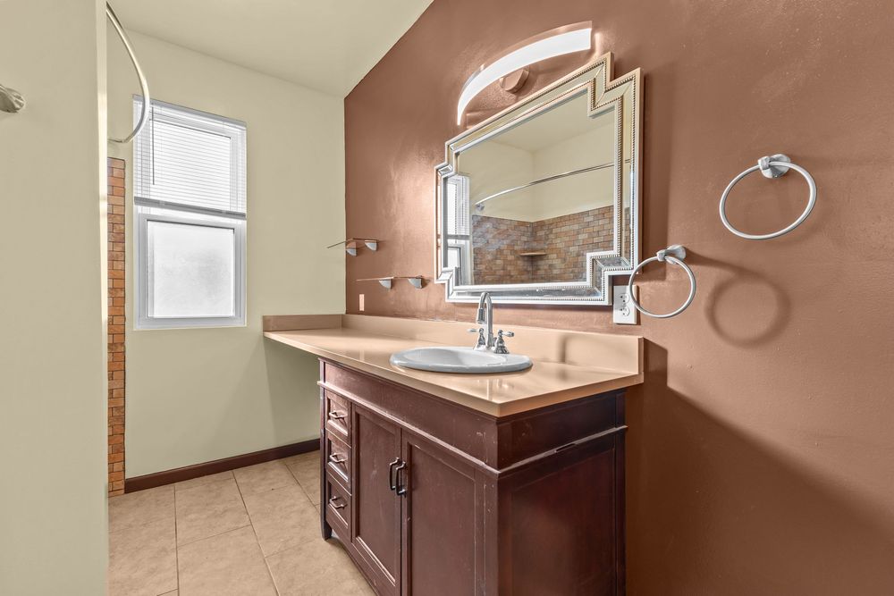 Bathroom vanity with dark wood cabinetry, decorative mirror with lighting, and neutral tile flooring.