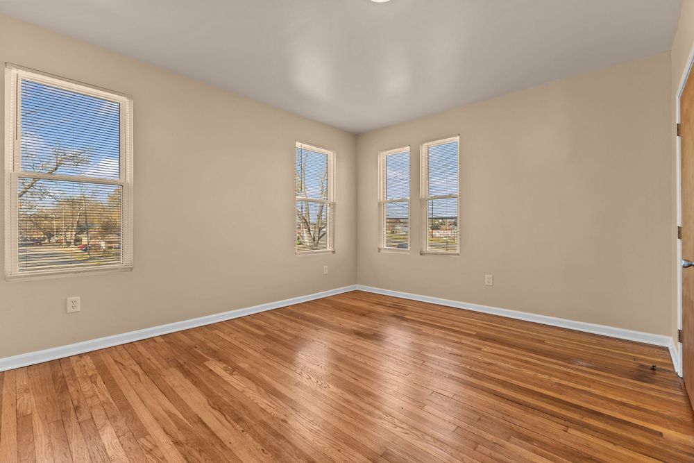 Empty bedroom with gleaming hardwood floors, neutral walls, and two bright windows with natural light.