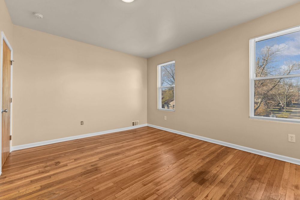 Bright empty bedroom with hardwood floors, neutral walls, and two windows with natural light.