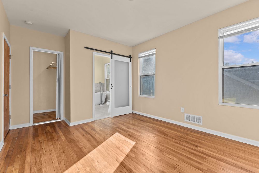 Empty bedroom with hardwood floors, barn door, closet, and bright window with natural light.