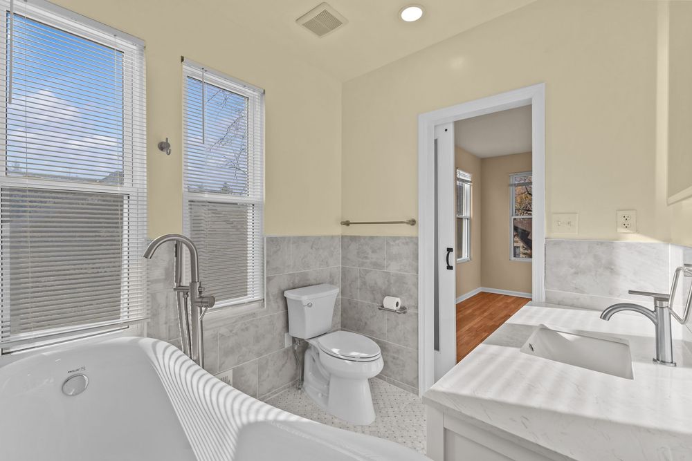 Spacious bathroom with soaking tub, gray tile walls, double vanity, and bright windows.
