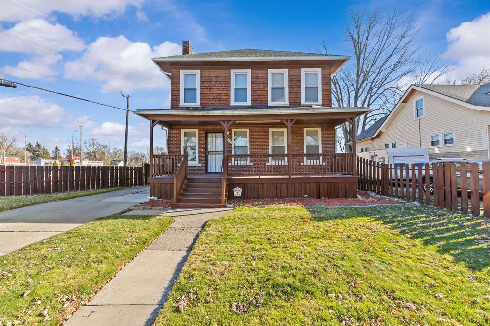 Brick two-story home with covered front porch, wood deck, and fenced yard with green lawn.