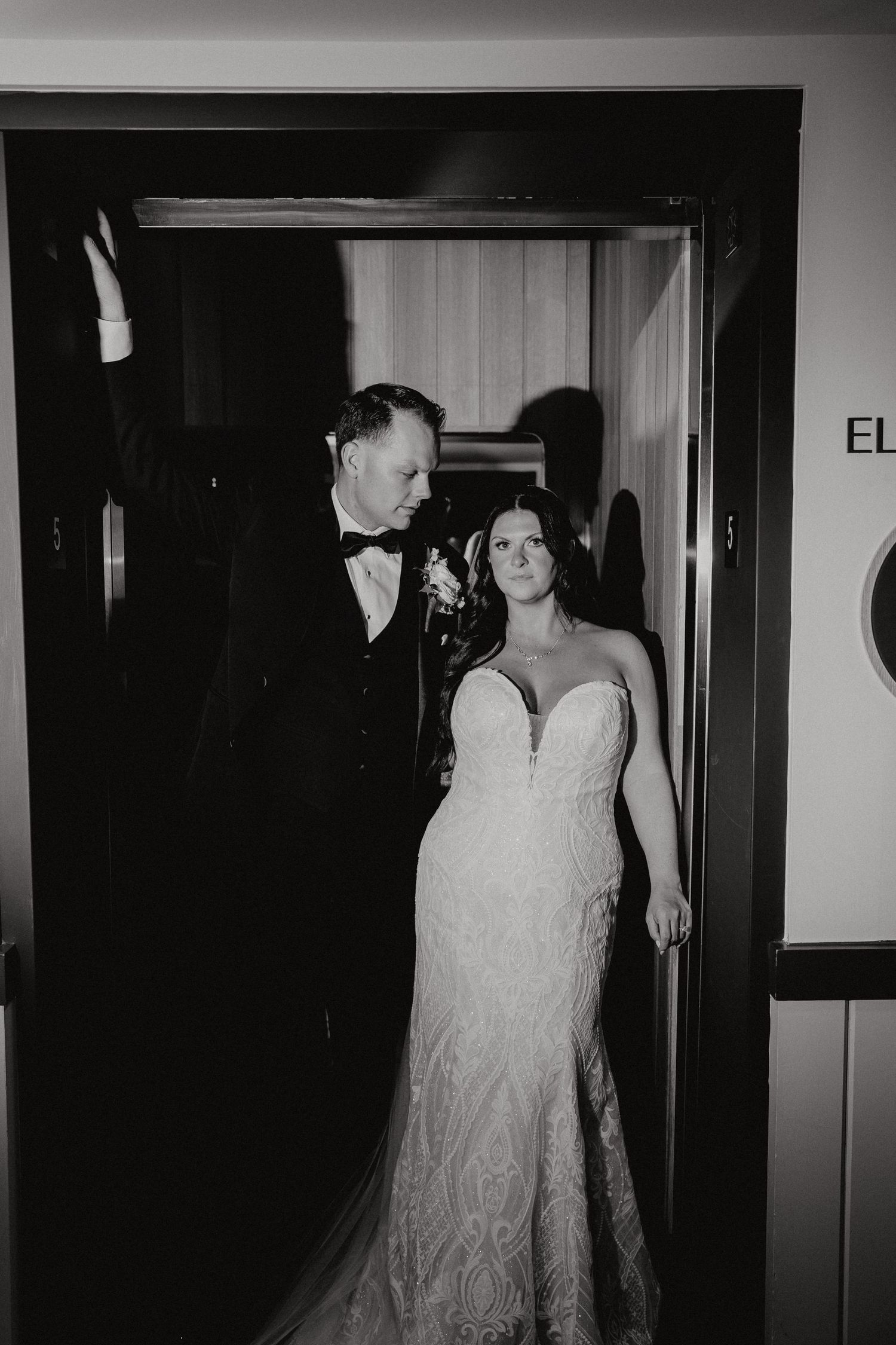 A couple stands in a doorway at their wedding, with the bride wearing a fitted lace gown and the groom in a black tuxedo.
