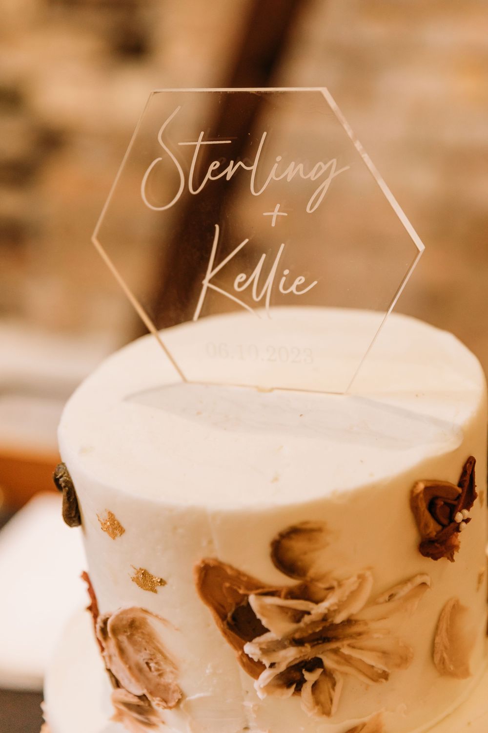 A white wedding cake decorated with brown floral designs and a clear hexagonal acrylic topper with elegant script text.