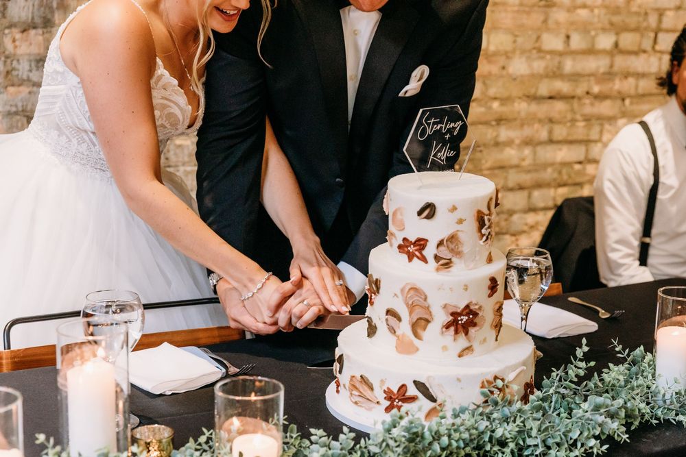 Hands cutting into an elegant white wedding cake decorated with delicate copper-colored autumn leaves.