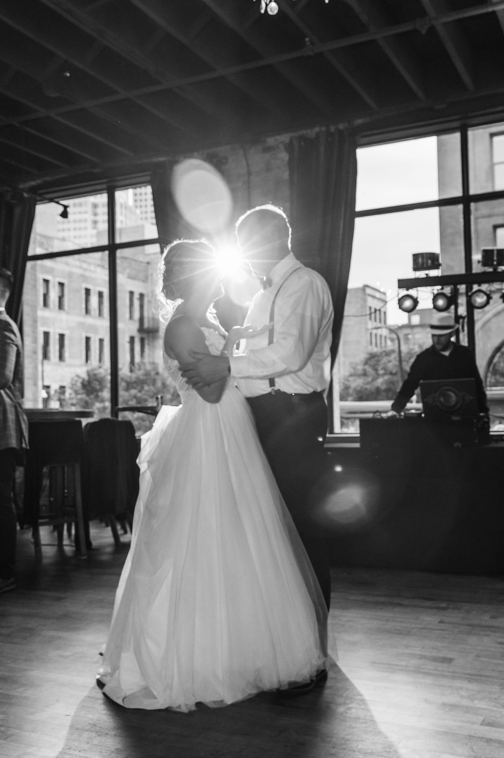 A couple shares their first dance in an elegant ballroom as sunlight streams through large windows.