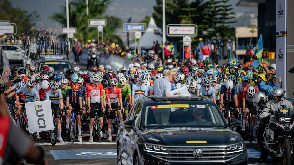 A large group of cyclists ride through city streets during a cycling event with support vehicles following behind.