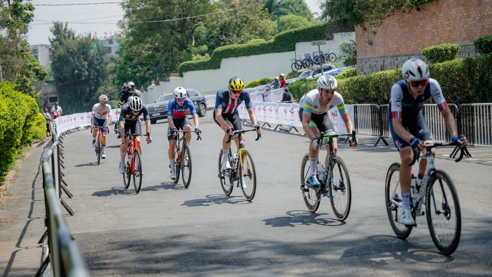Cyclists race around a corner during a competitive road cycling event on a sunny day.
