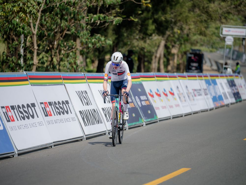 Cyclist racing down a paved road with advertising banners and barriers along the course during a competition event.