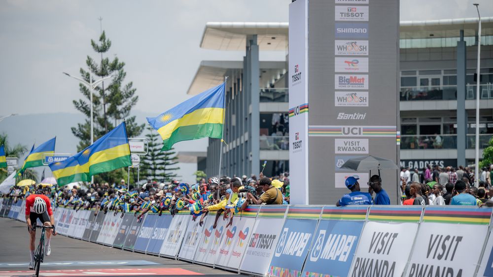 Crowd gathers at outdoor sporting event with flags and barriers visible near a large building structure.