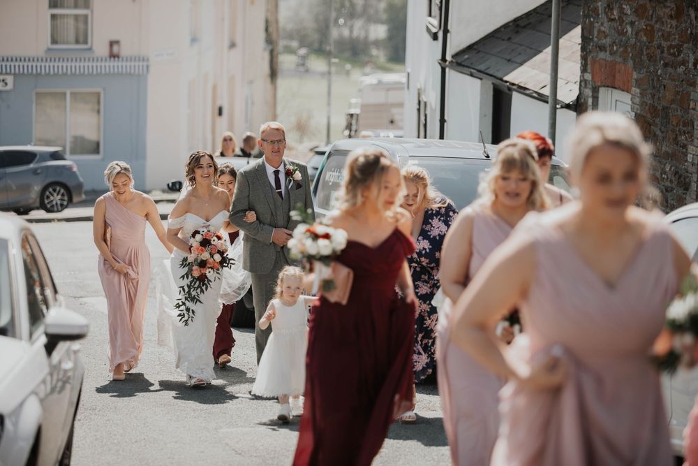 Wedding party walking down a sunny street, bride in white gown with bridesmaids in blush and burgundy dresses.
