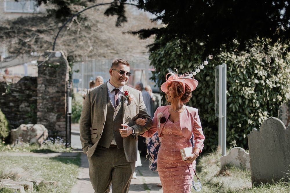 Elegantly dressed wedding guests walking through a churchyard, man in tweed suit and woman in pink fascinator hat.