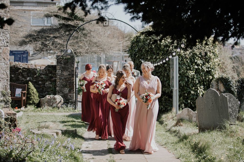 Bridesmaids in burgundy and blush gowns holding bouquets walking through a sunlit church garden path.