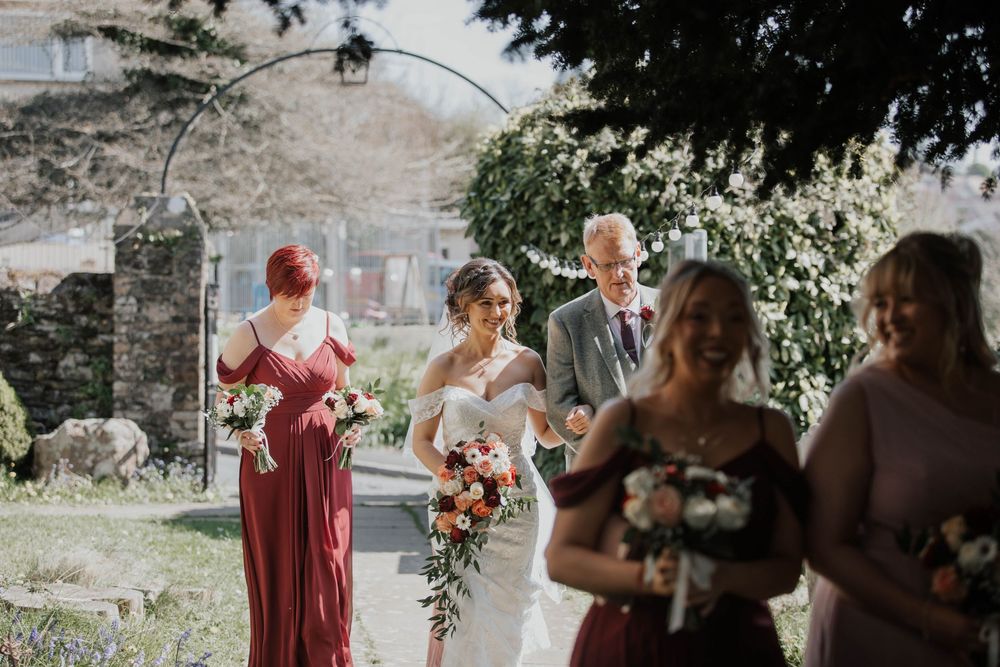 Bride in white gown walking with groom and bridesmaids in burgundy dresses through a sunny church garden.