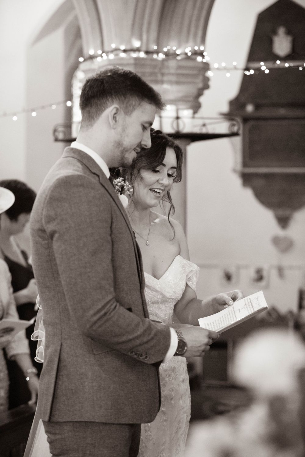Black and white photo of bride and groom sharing vows in a church decorated with fairy lights.