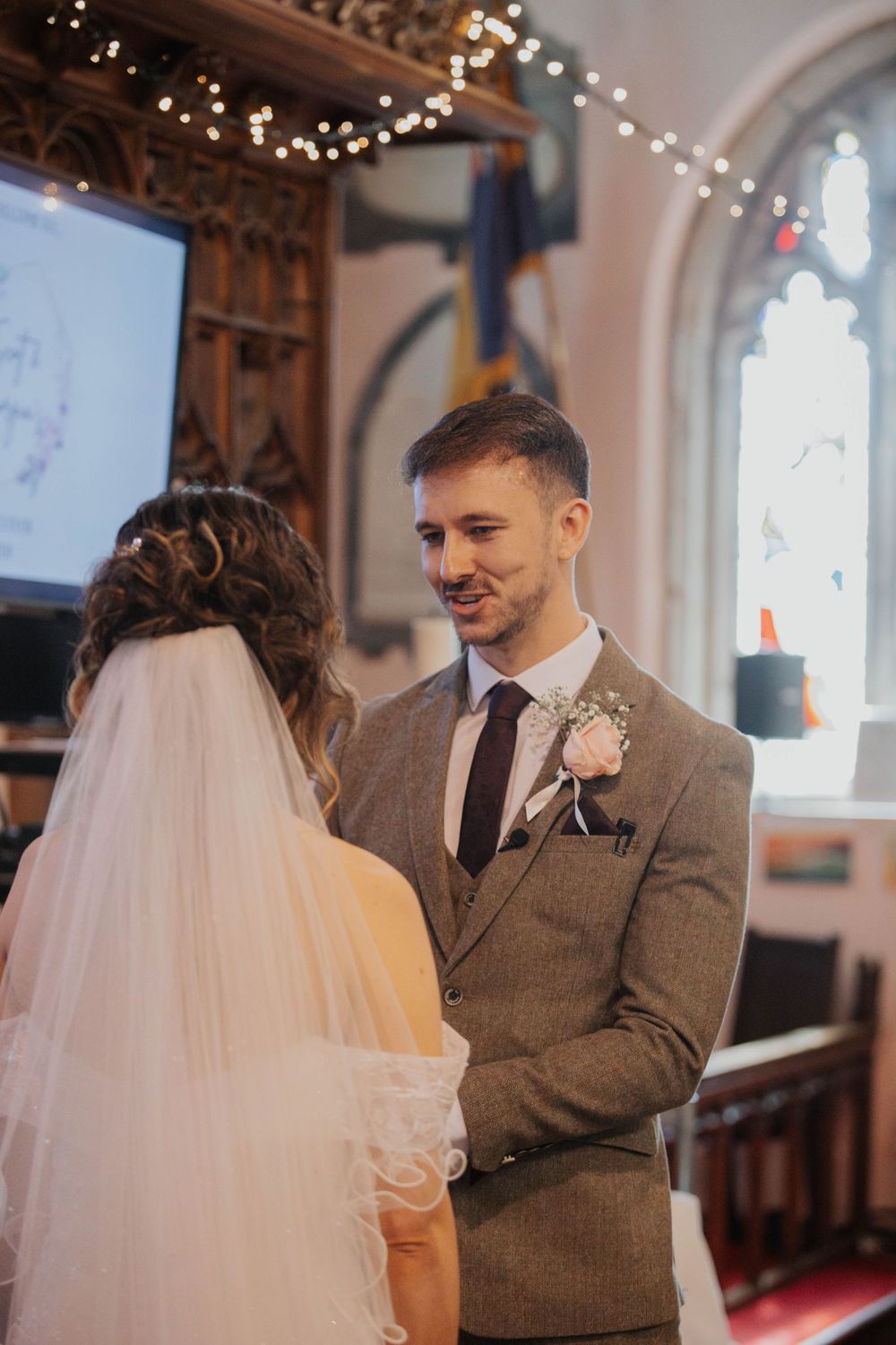 Smiling groom in grey tweed suit with boutonniere facing bride during church wedding ceremony.