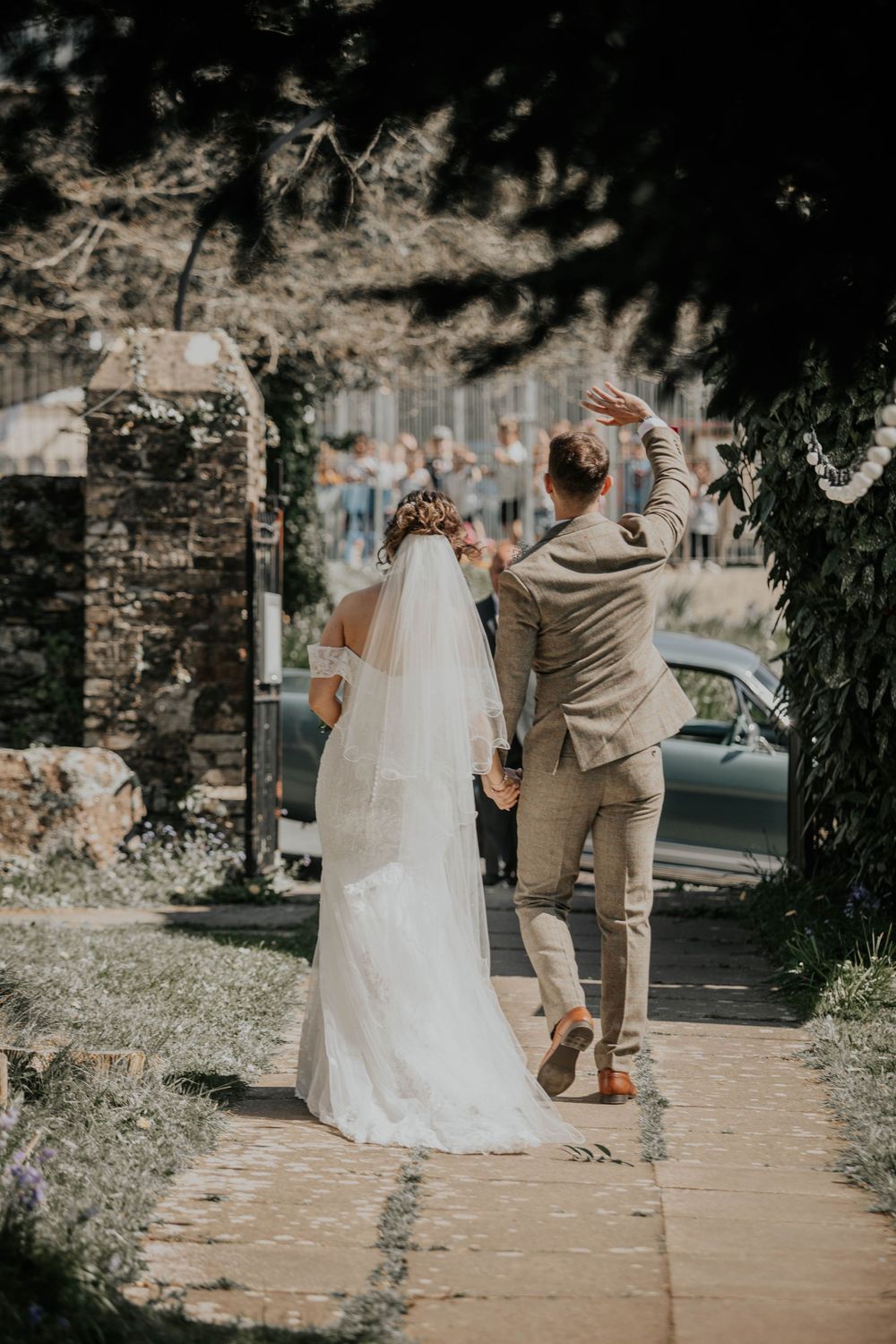 Bride and groom walking away down a garden path, groom waving to wedding guests ahead.