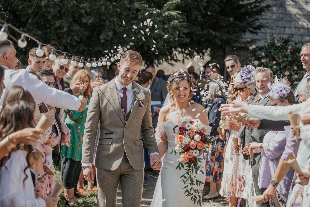 Bride and groom walk through confetti shower surrounded by cheering wedding guests outdoors.