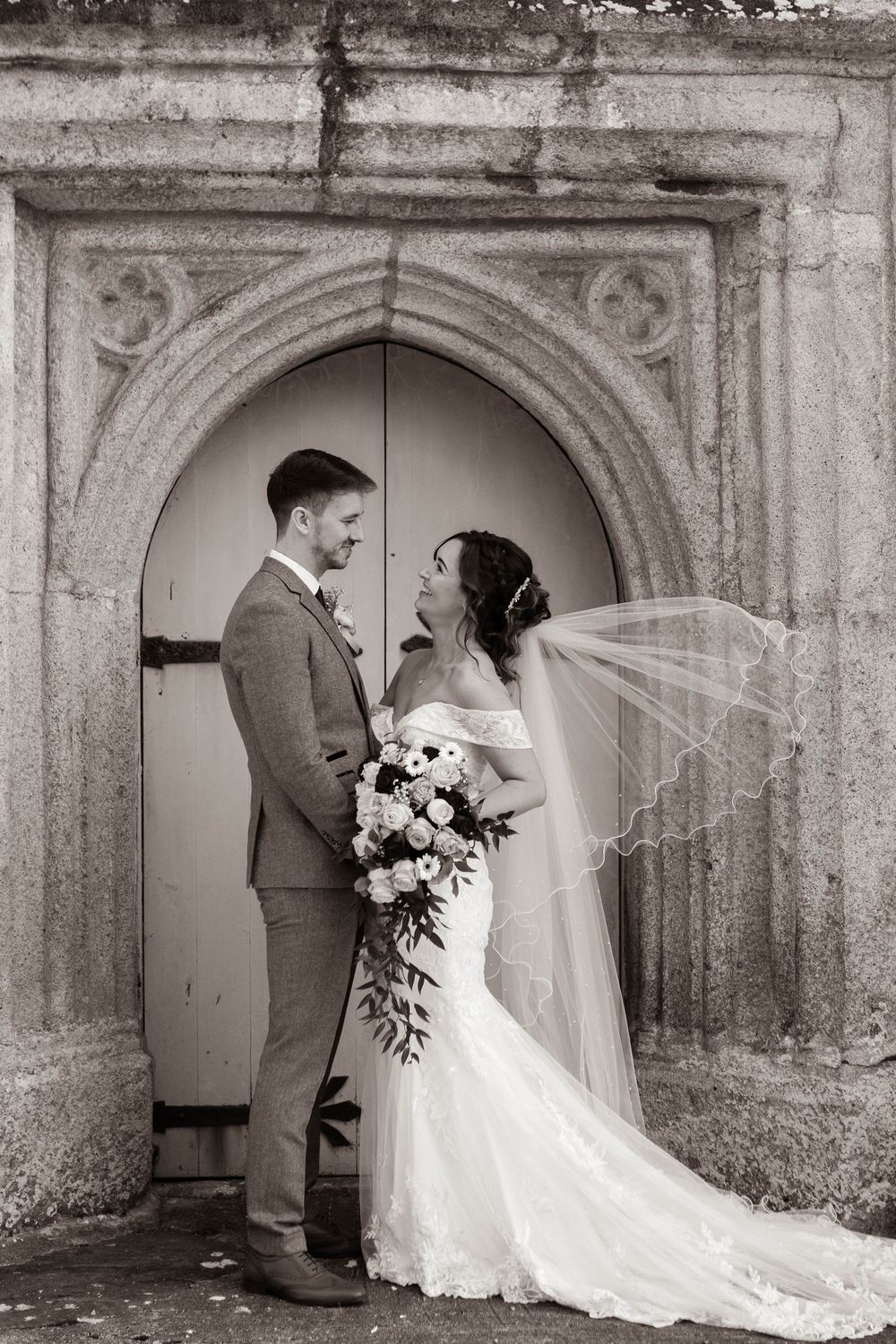 Black and white portrait of bride and groom holding hands before ornate church doorway.