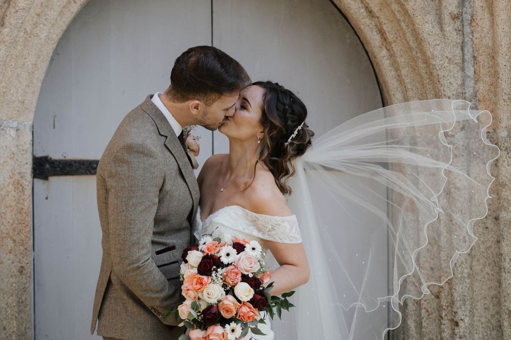 Bride and groom share a romantic kiss outside a stone church doorway with veil flowing.