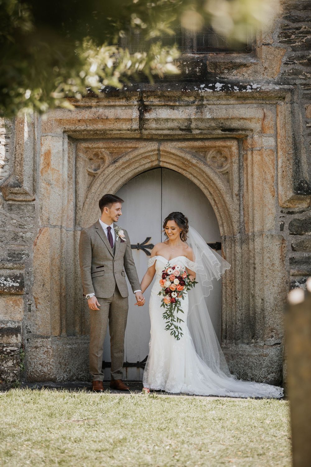 Bride and groom hold hands smiling before a grand stone arched church doorway outdoors.