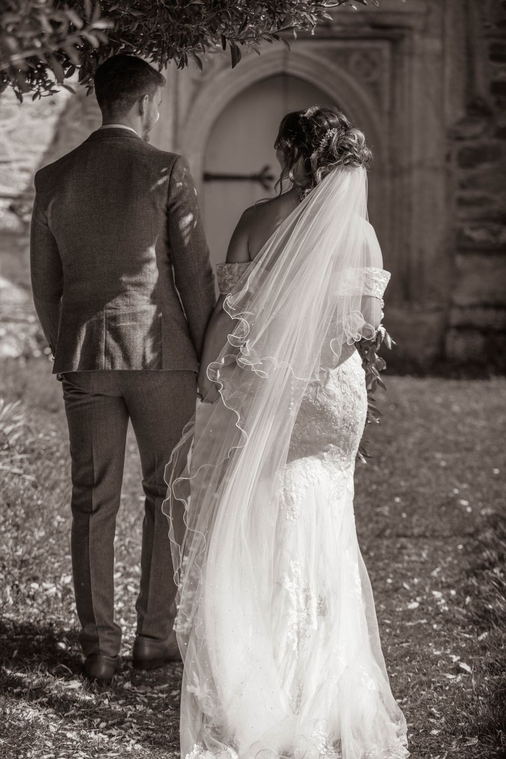 Black and white rear view of bride and groom standing together outside historic stone church.