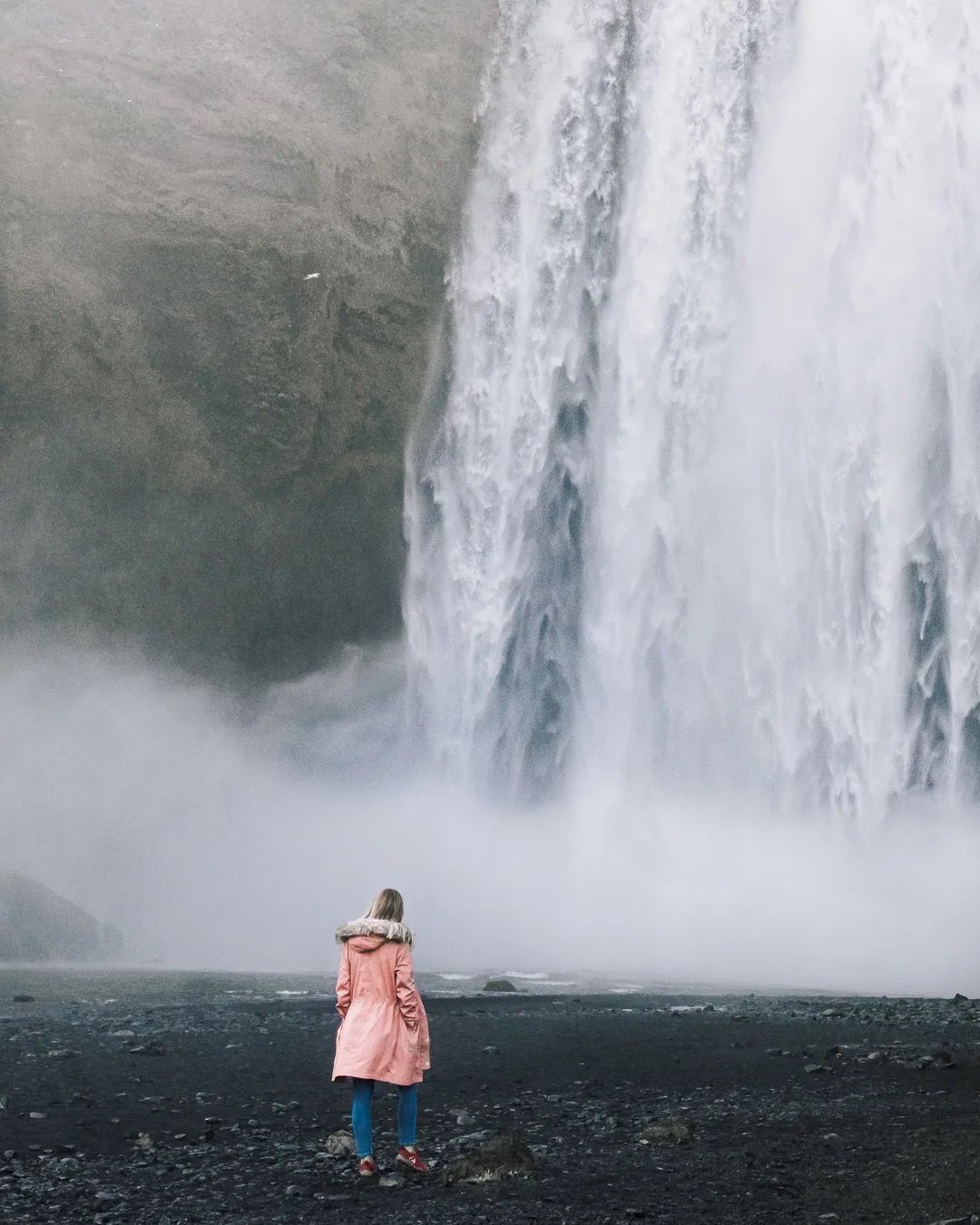 Woman in a pink coat standing in mist at the base of a tall Icelandic waterfall.