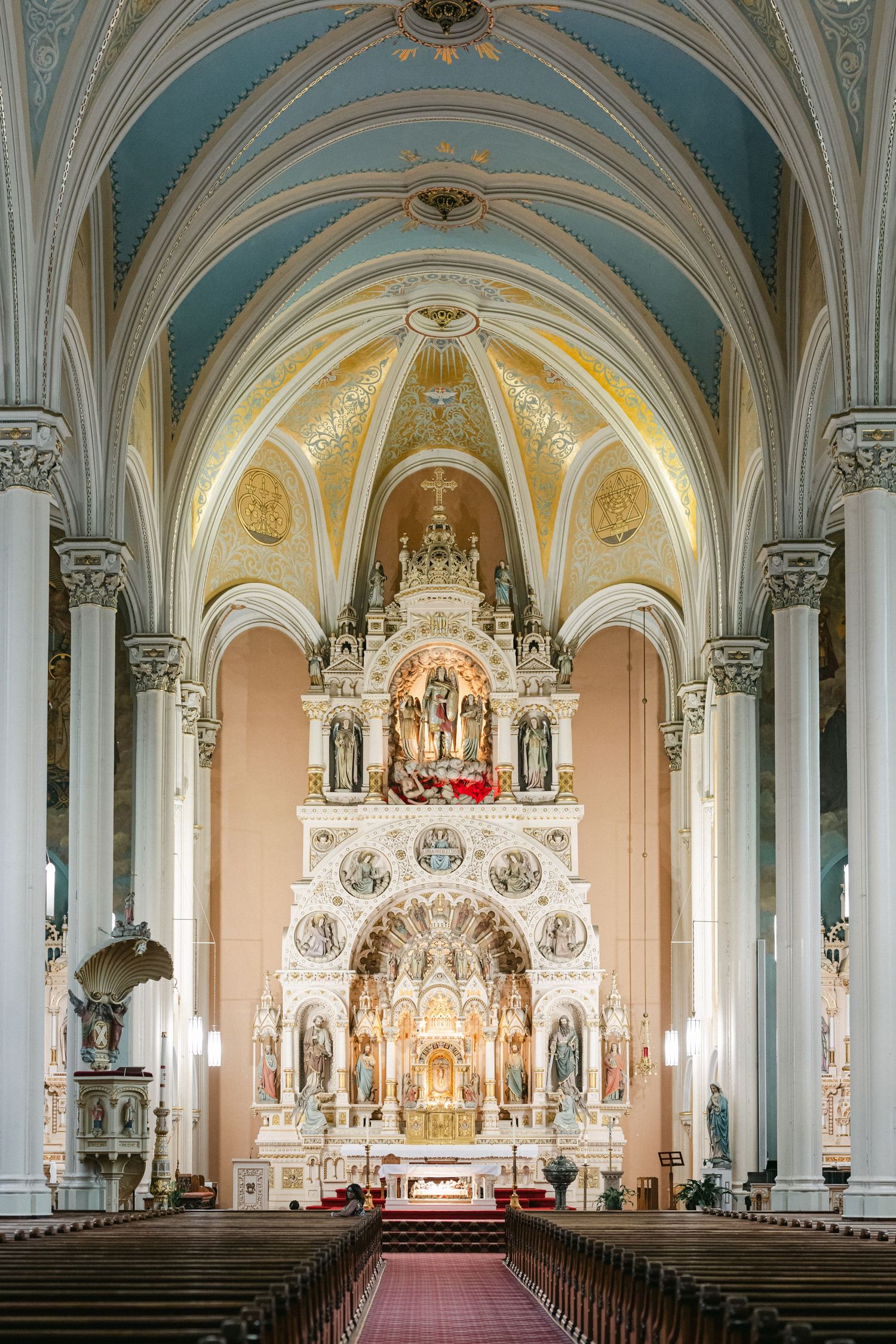 Interior panorama of a grand Catholic church with ornate altar, arched ceilings, and crystal chandeliers.