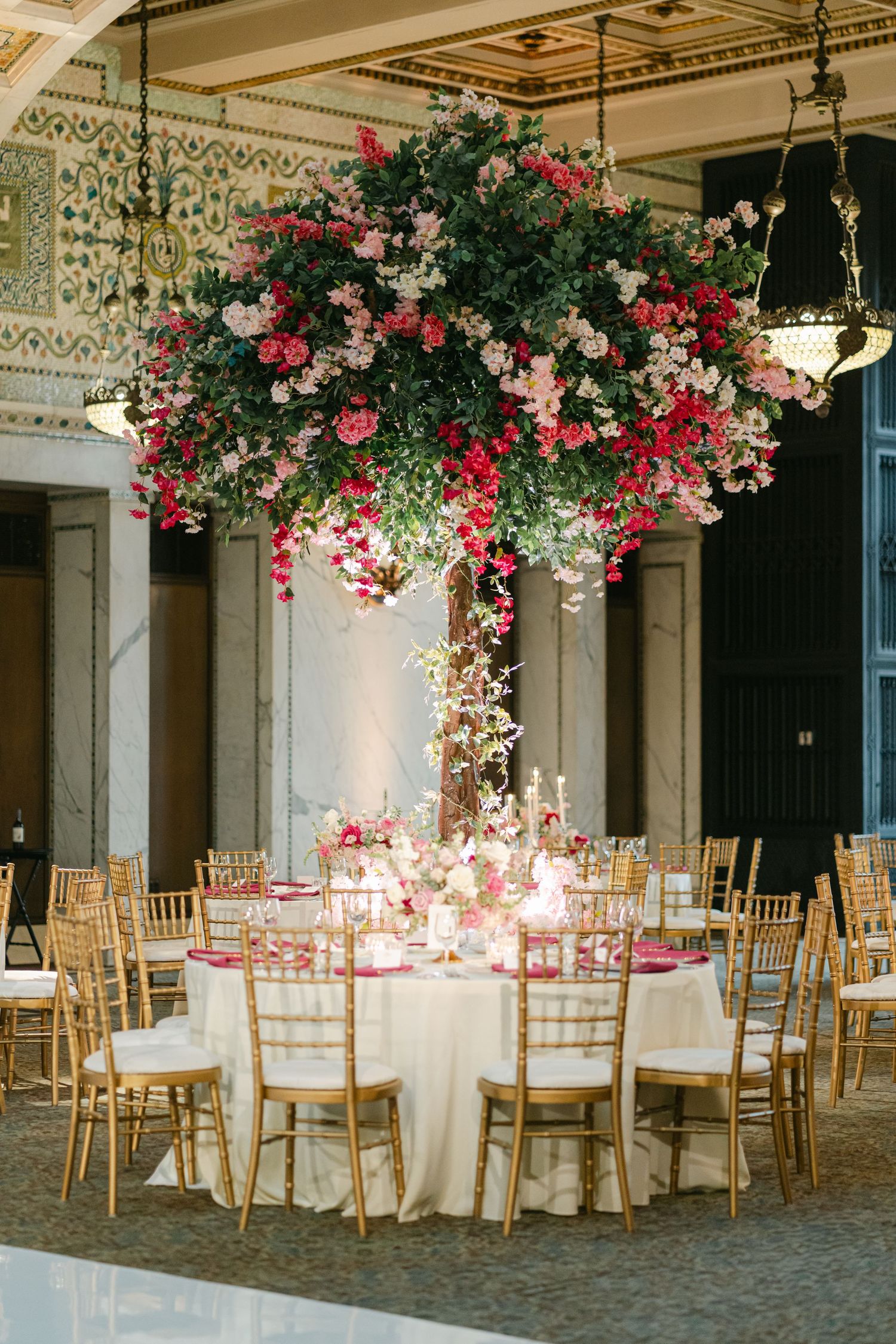 Elegant wedding reception venue with hanging floral arrangements, gold chiavari chairs, and round tables with white linens.