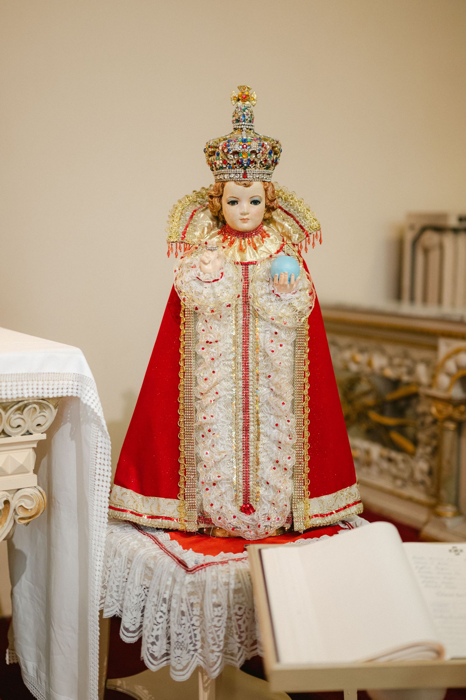 Ornate infant Jesus religious dress with white lace, red velvet cape, and golden crown displayed on white furniture.
