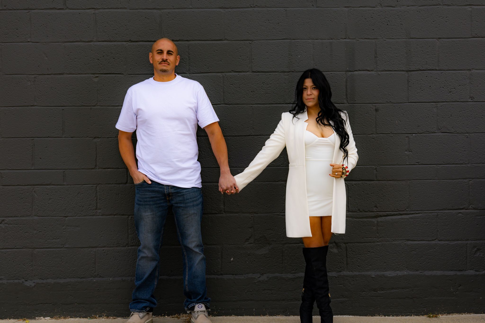 A couple poses together against a dark wall in casual attire.