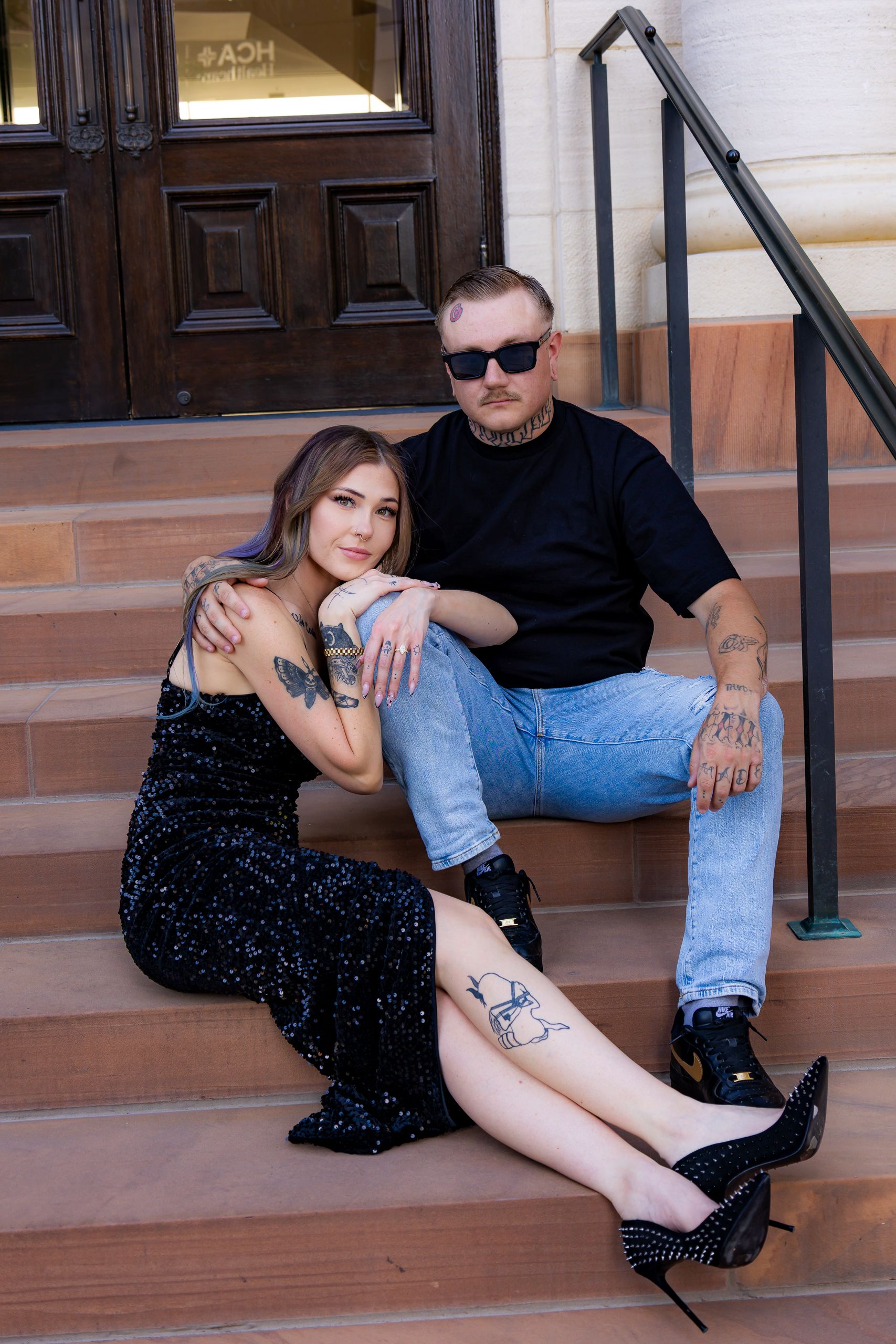 Tattooed couple posing on stone steps outside a building, woman in black sequin dress with heels, man in black tee and jeans.