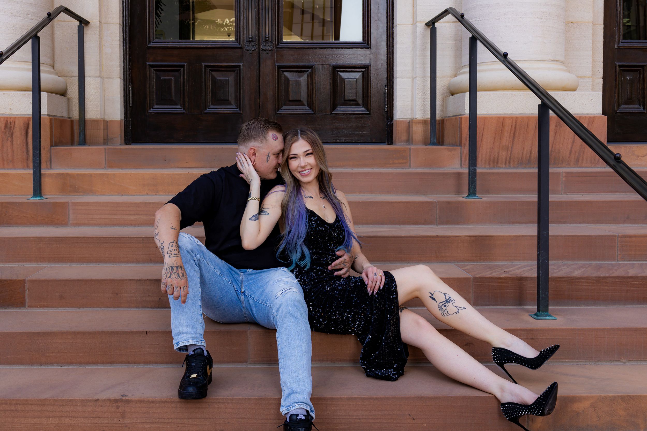 Couple sitting on brick steps outside a building, man whispering to woman in black dress with purple hair.