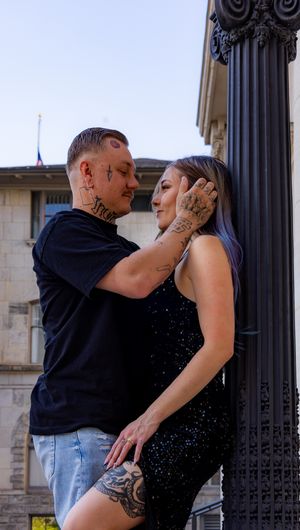 A tattooed couple in black outfits share an intimate moment by a decorative pillar outdoors.
