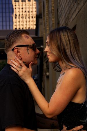 A tattooed couple shares an intimate moment outdoors at golden hour, with the woman gently holding the man's face.