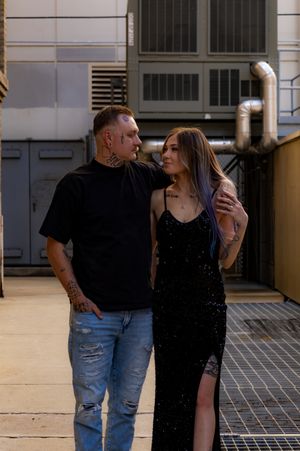Tattooed couple posing together in industrial setting, man in black tee and ripped jeans, woman in black sequin dress.