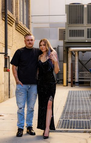 A tattooed man in a black tee and jeans poses with a woman in a sparkly black gown in an urban alley.