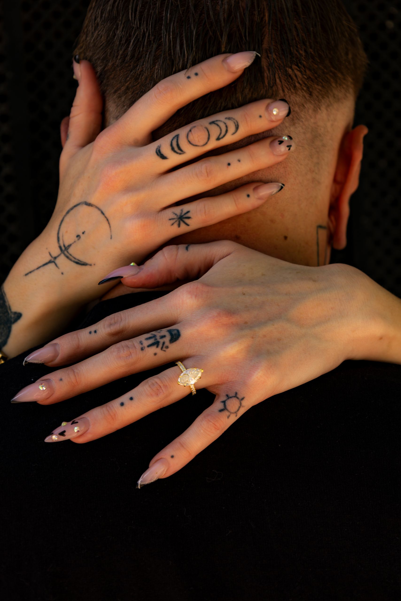 Two hands with celestial finger tattoos including moons, stars, and suns rest on a person's neck, with long nude stiletto nails.