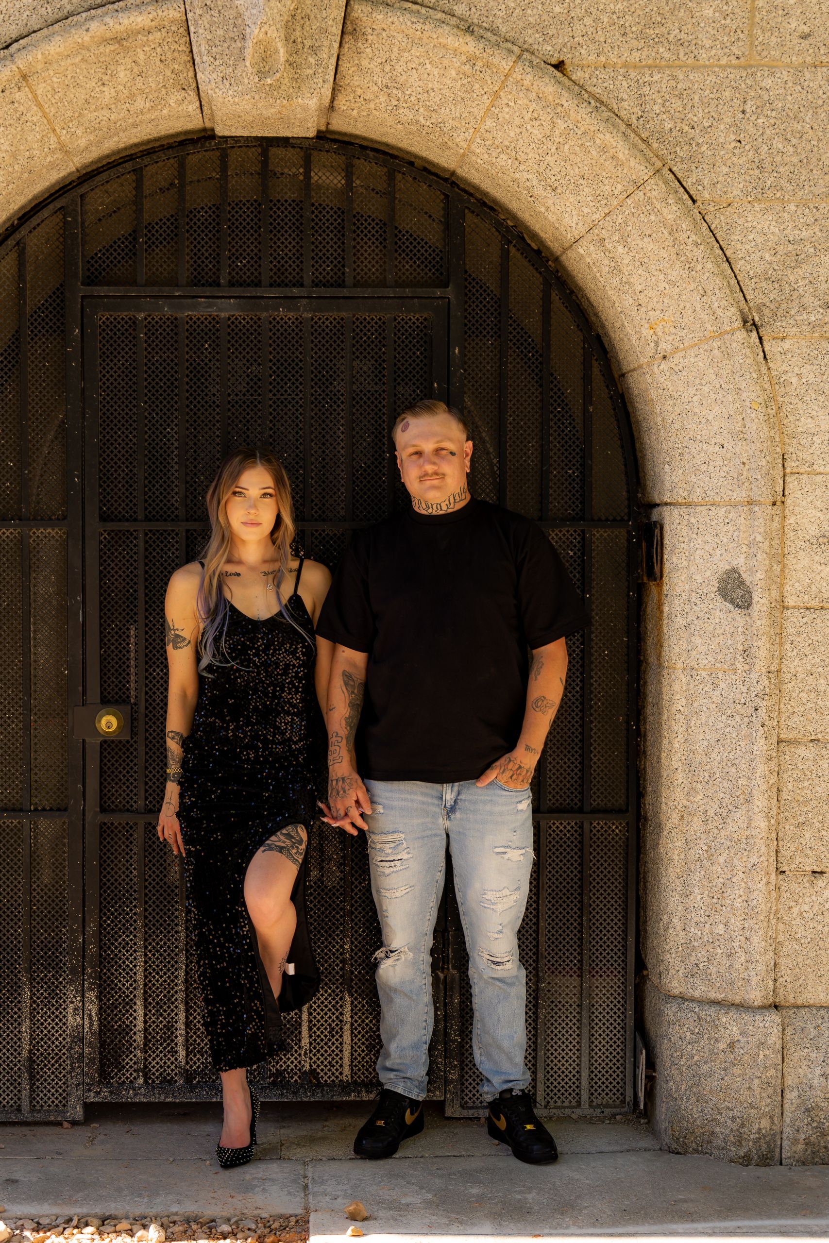 A couple poses in front of an arched stone doorway, woman in black dress, man in black tee and jeans.
