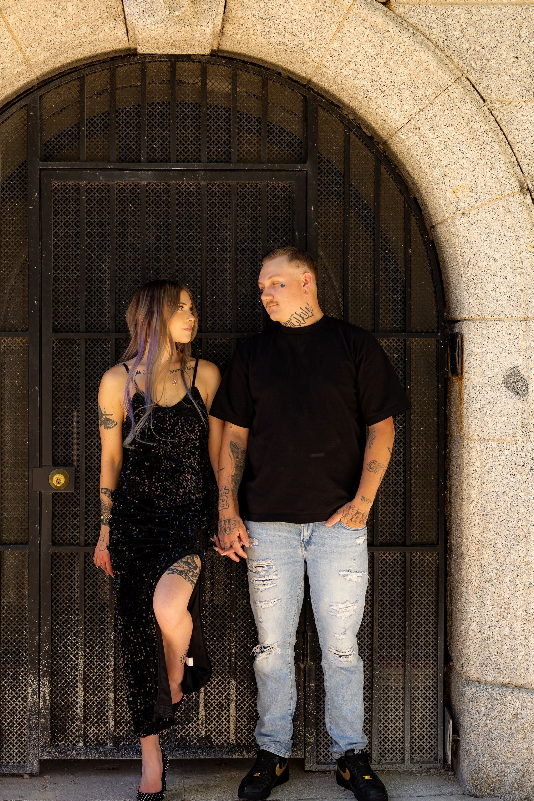 A tattooed couple holds hands standing before an arched stone doorway, wearing black outfits and jeans.