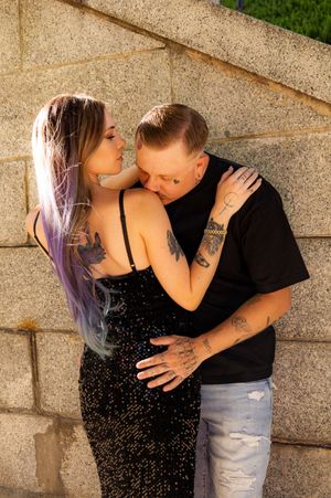 A couple with tattoos share an intimate embrace against a stone wall, the woman wearing a black sequin dress.