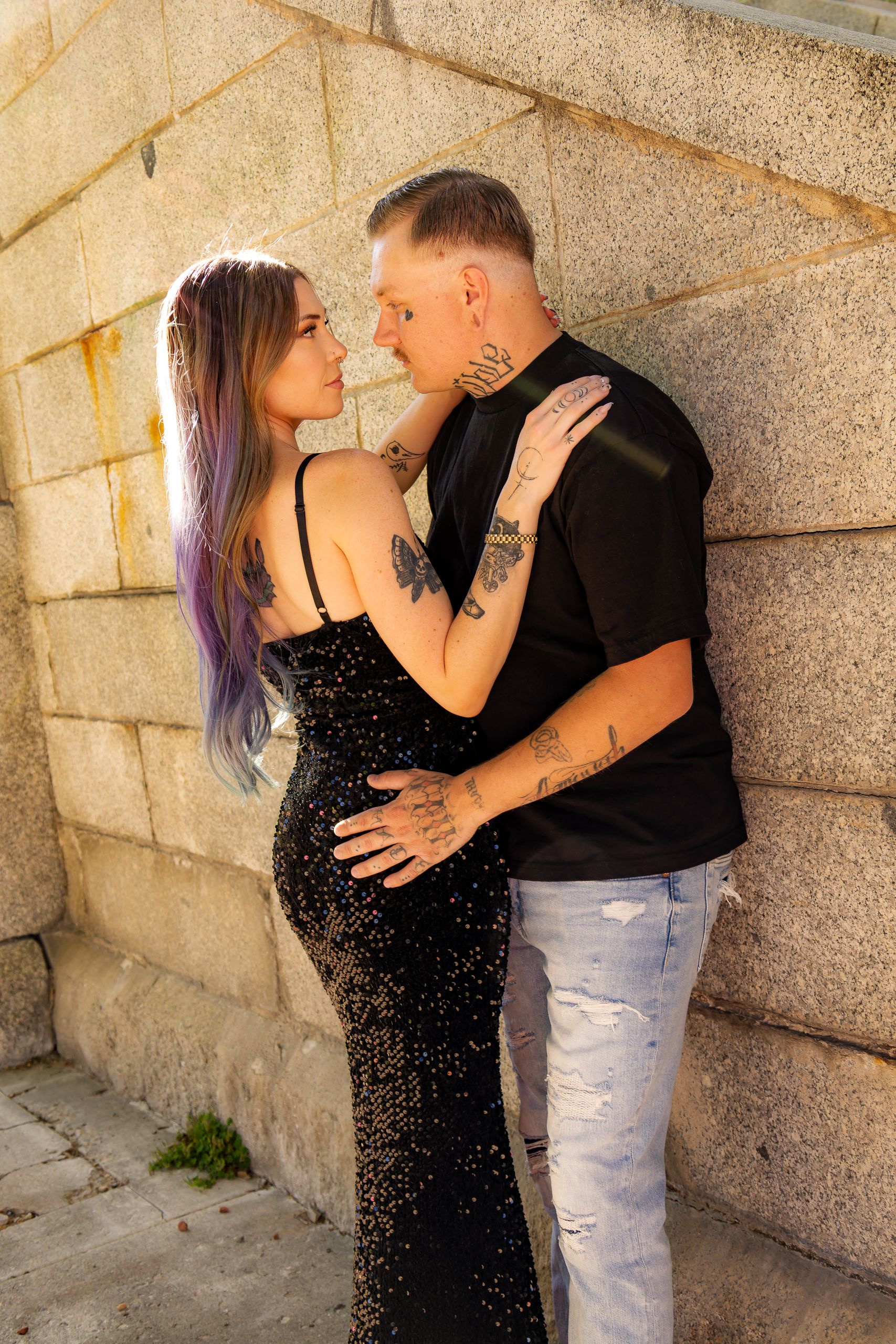 Tattooed couple in black outfits sharing a romantic moment against a stone wall, woman in sequin dress with purple hair.