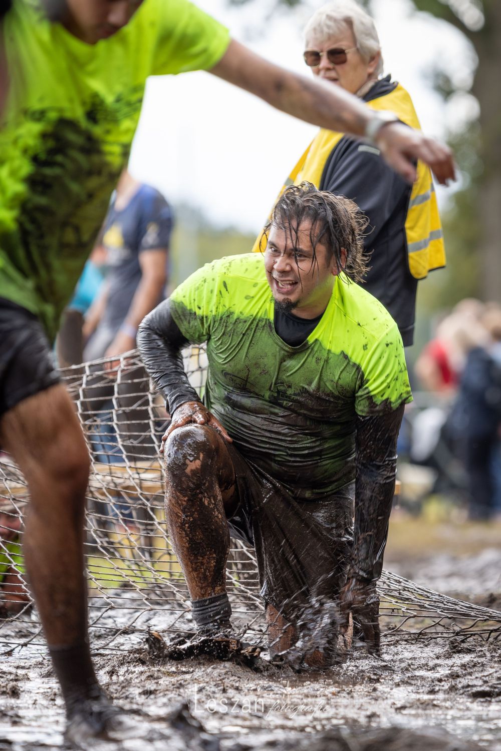 Participants crawl through a muddy obstacle course under metal wire fencing during an outdoor challenge event.