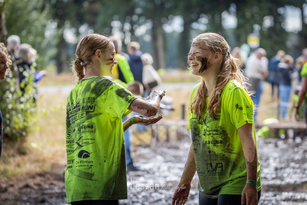 Two volunteers in bright yellow-green t-shirts laugh together while working at an outdoor community event in muddy conditions.