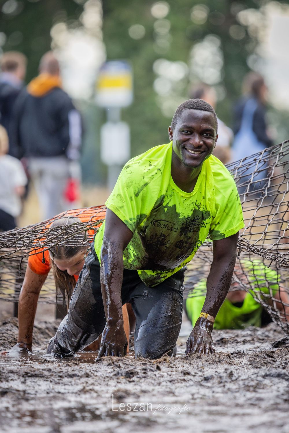 Athlete in bright yellow shirt crawls under wire mesh obstacle during muddy obstacle course race.
