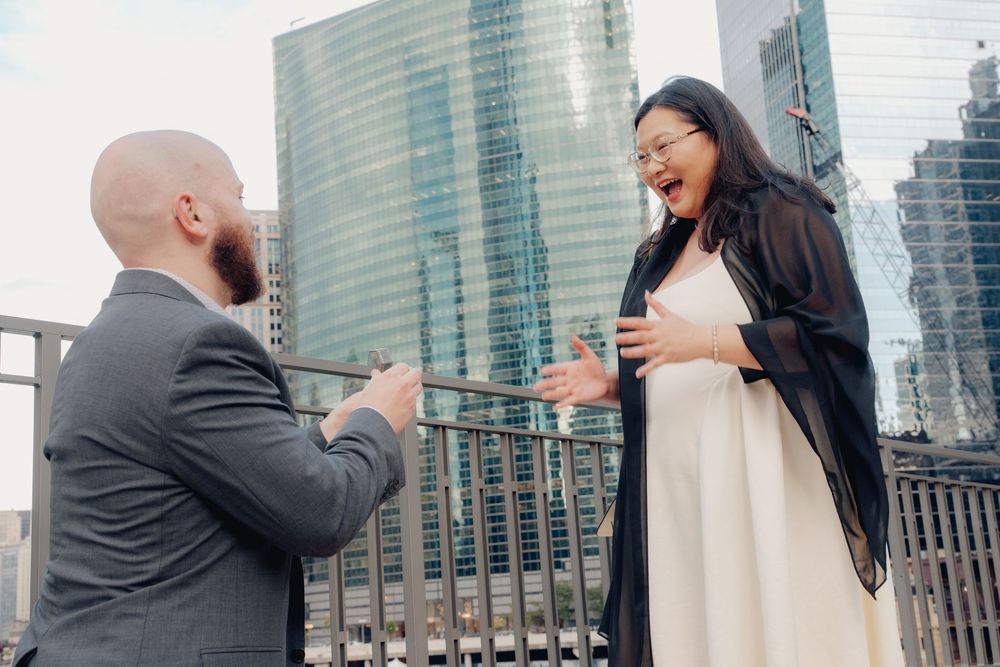 Man proposing to surprised woman on city rooftop with skyscrapers in background.