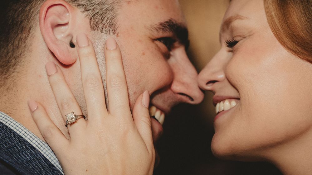 Close-up shot of a romantic couple smiling and touching faces with engagement ring visible on hand.